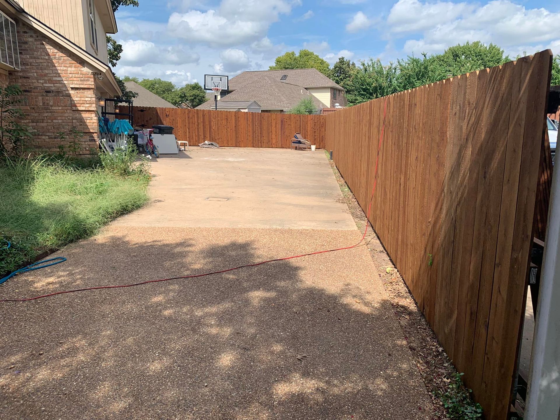 Gravel driveway with a wooden fence. Green grass and a house on the left. Sunny day.