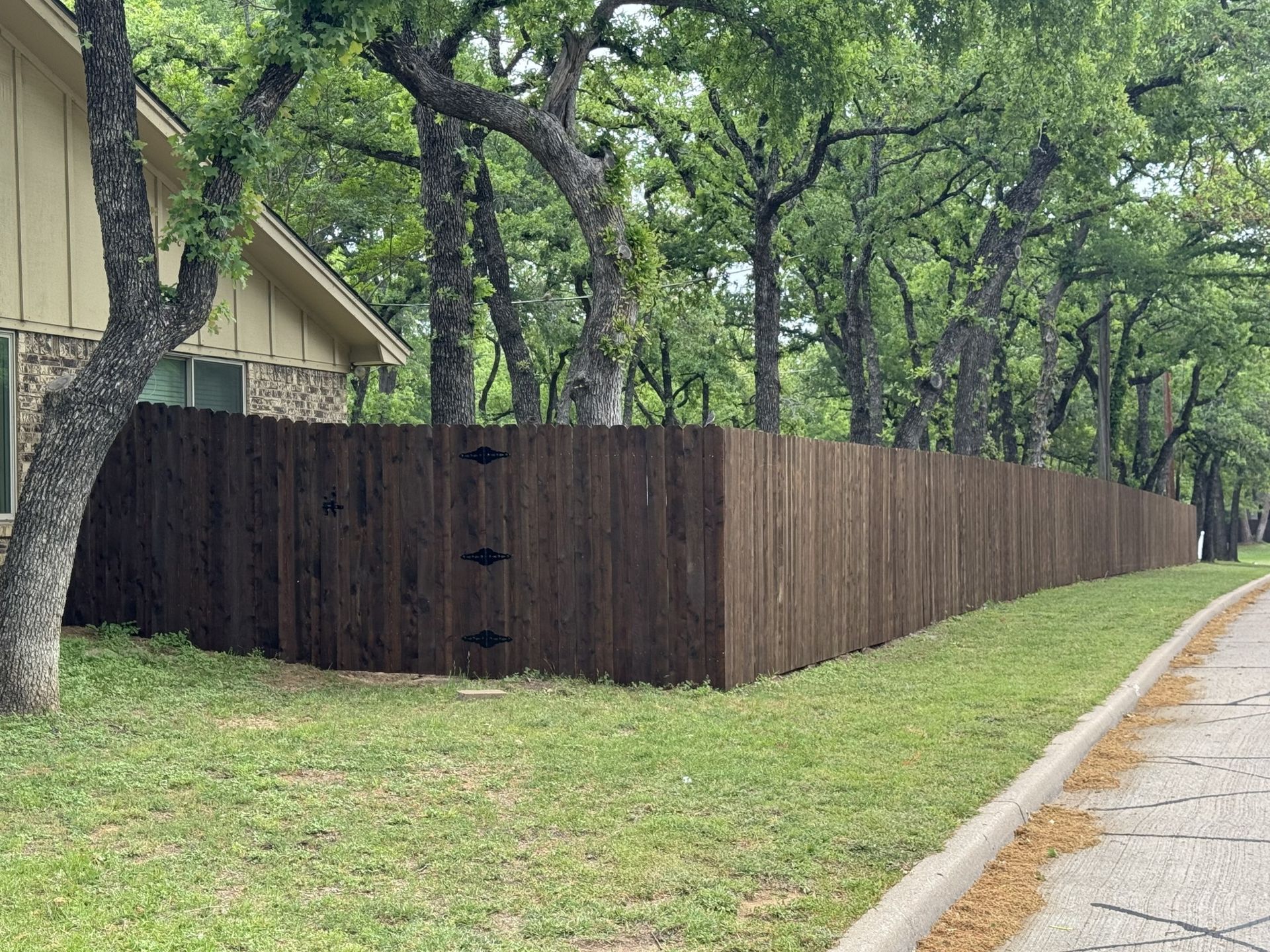 Brown wooden fence along a road lined with trees and grass, next to a tan house.
