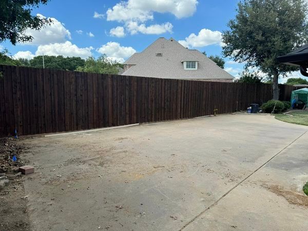 Brown wooden fence in a backyard, with a driveway and house in the background under a cloudy sky.