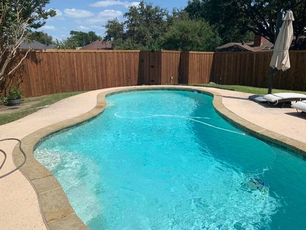 A backyard pool with clear blue water surrounded by concrete, a wooden fence, and lawn furniture on a sunny day.