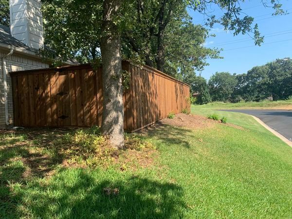 Brown wooden fence next to tree and a grassy hill with a paved road in the distance.