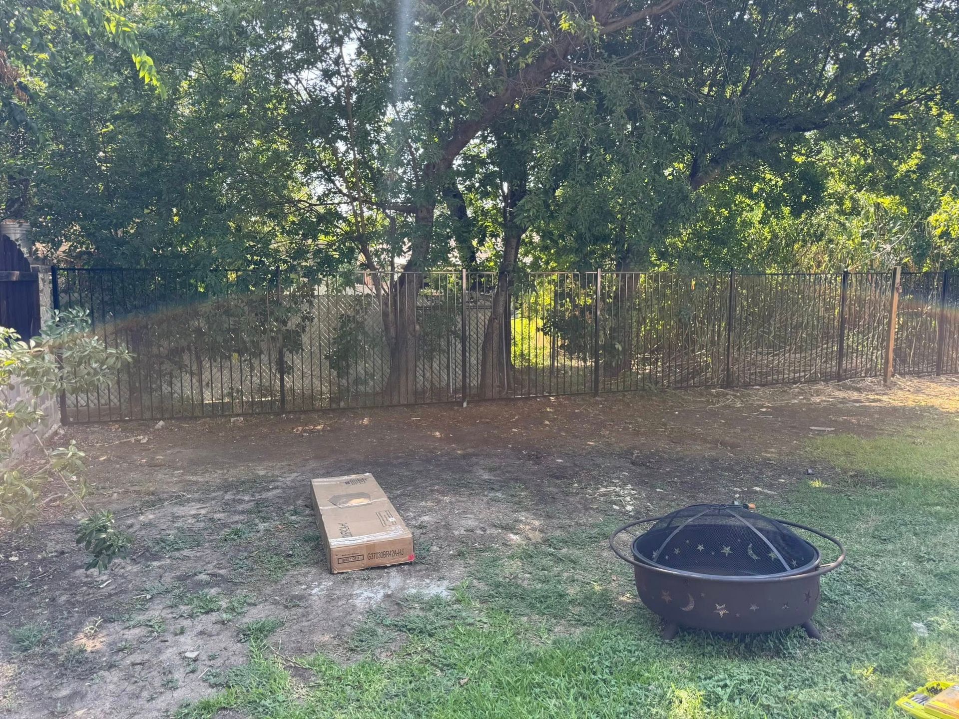 Backyard scene: metal fence, trees, fire pit, and a cornhole set.