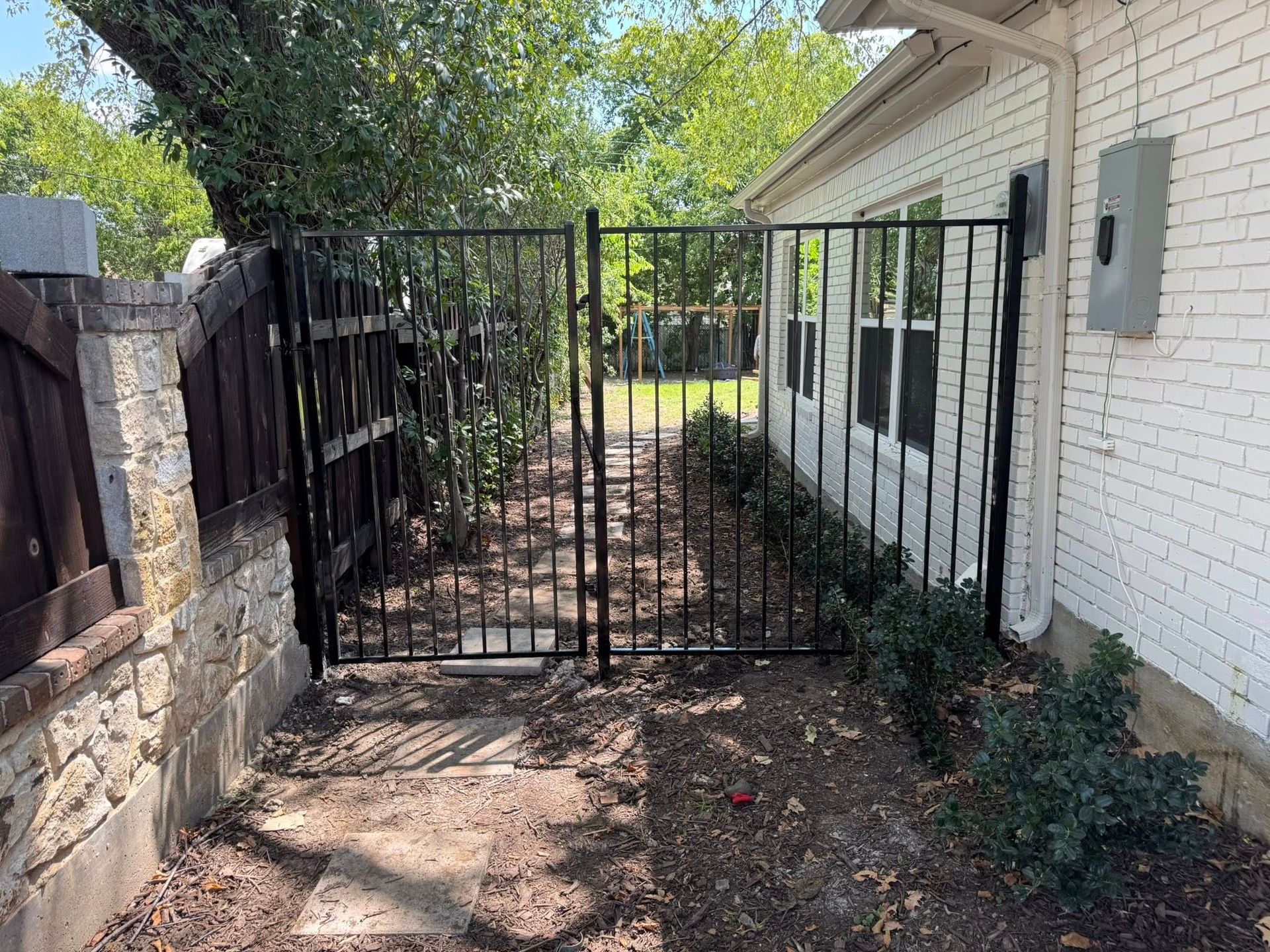 Black metal gate between a brick wall and a white house, leading to a yard.