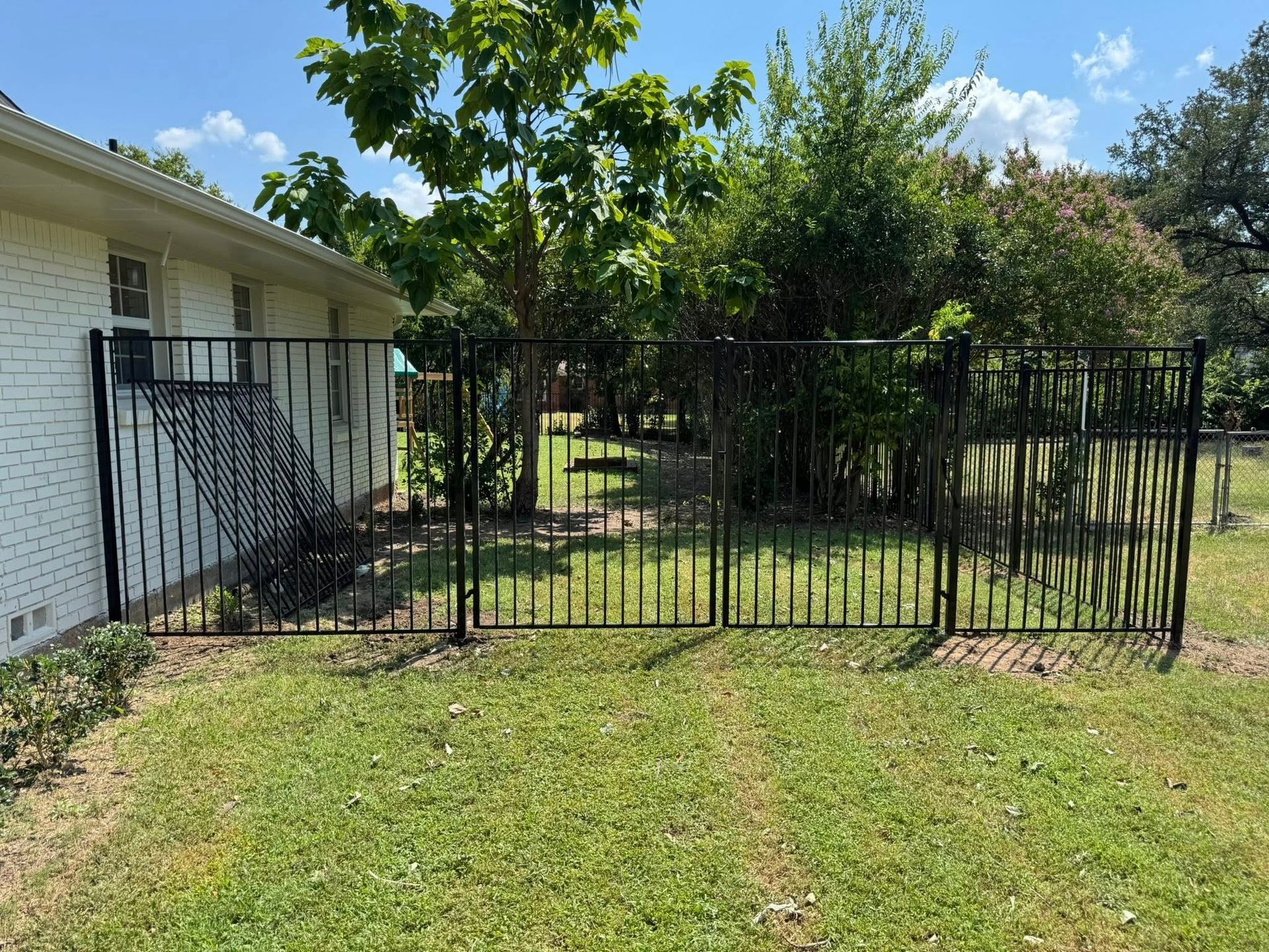 Black metal fence enclosing a grassy yard, next to a brick house and trees.