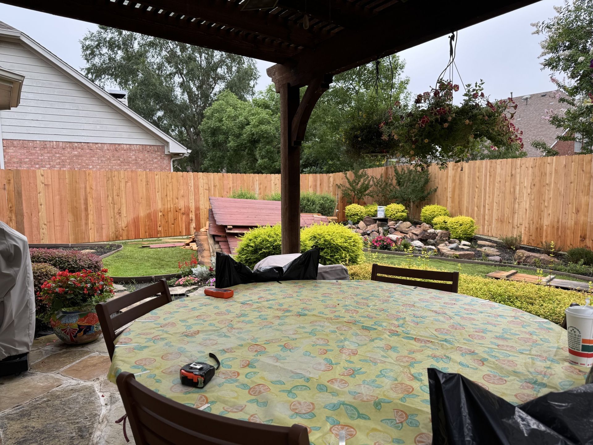 Backyard patio with a table, chairs, a wooden fence, and lush greenery under a pergola.