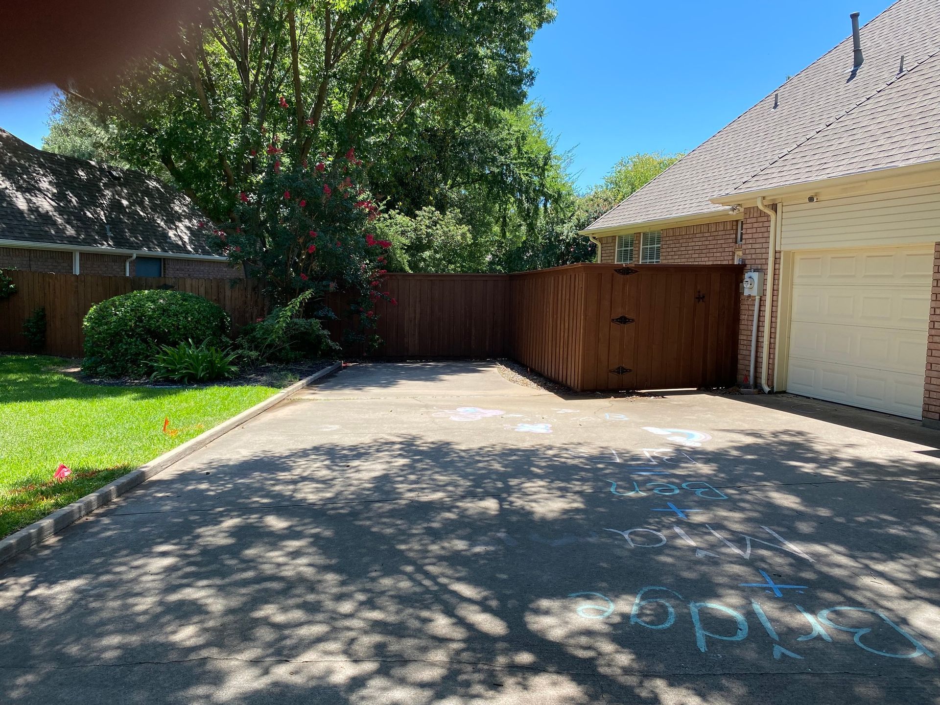 Driveway with brown fence, garage, green grass, and trees under a sunny blue sky.