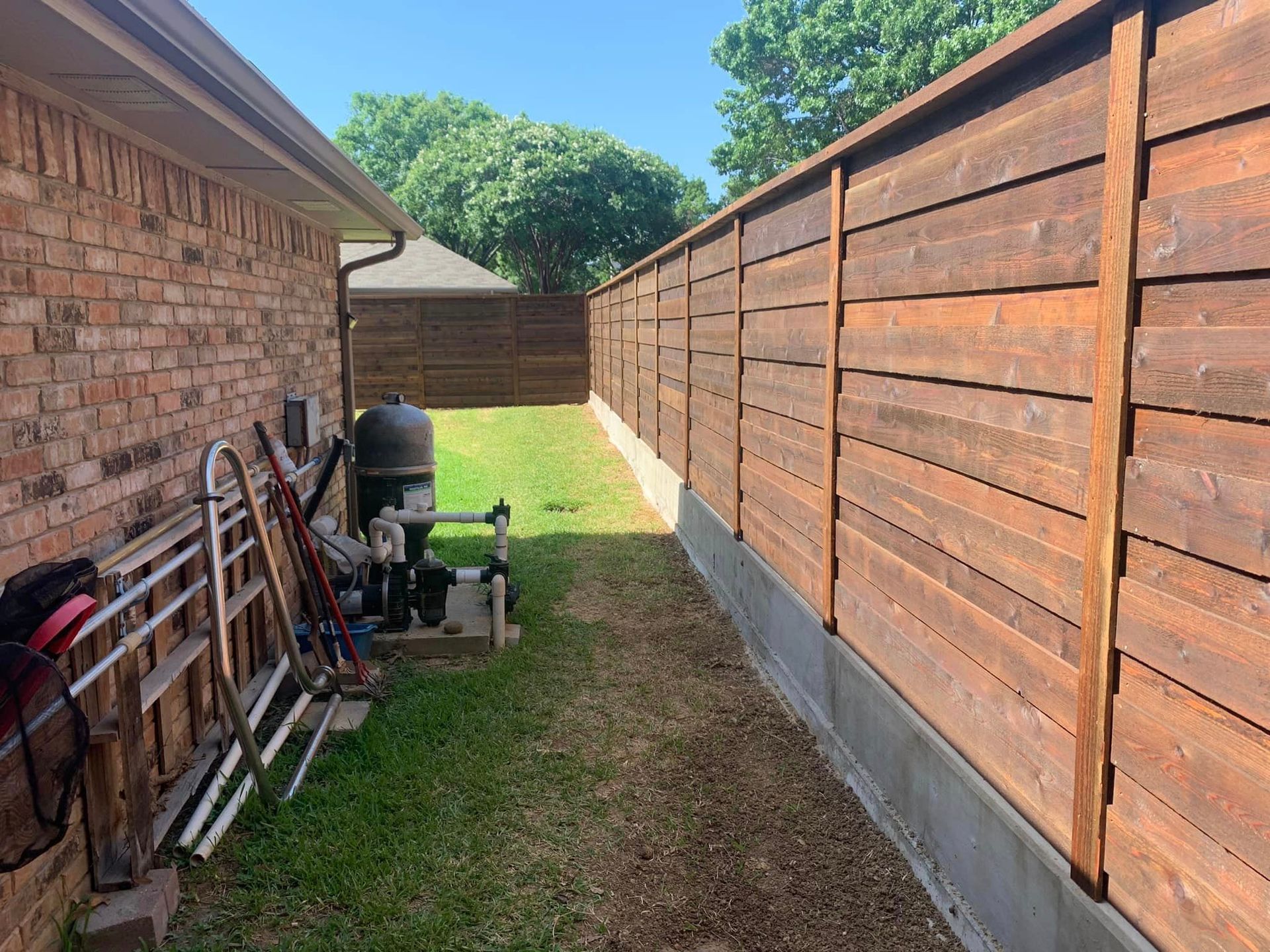 Backyard with a wooden fence, brick house, and lawn. Equipment near the house.