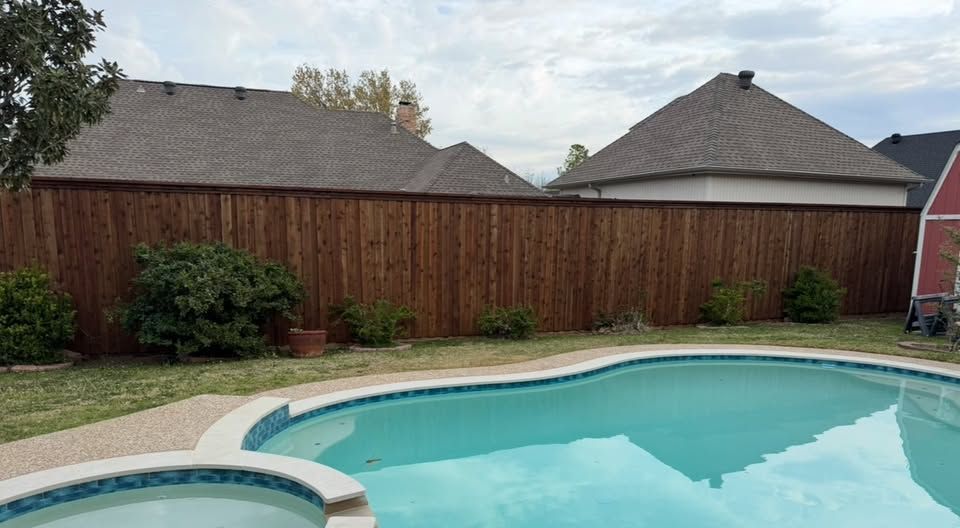 A backyard with a pool, wooden fence, grass, and houses under a cloudy sky.