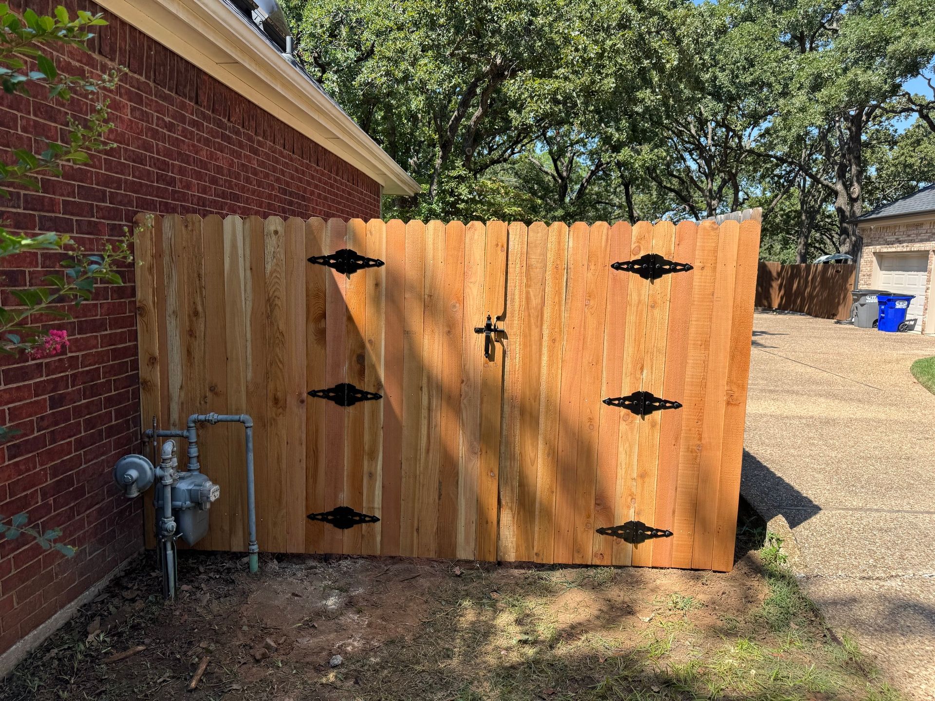Wooden gate with black hinges and latch in front of a brick building, covering utility meters.