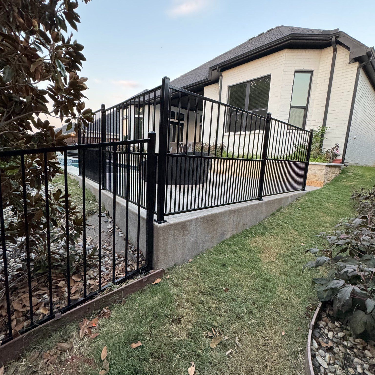 Black fence surrounds a patio with a house in the background and green grass in front.