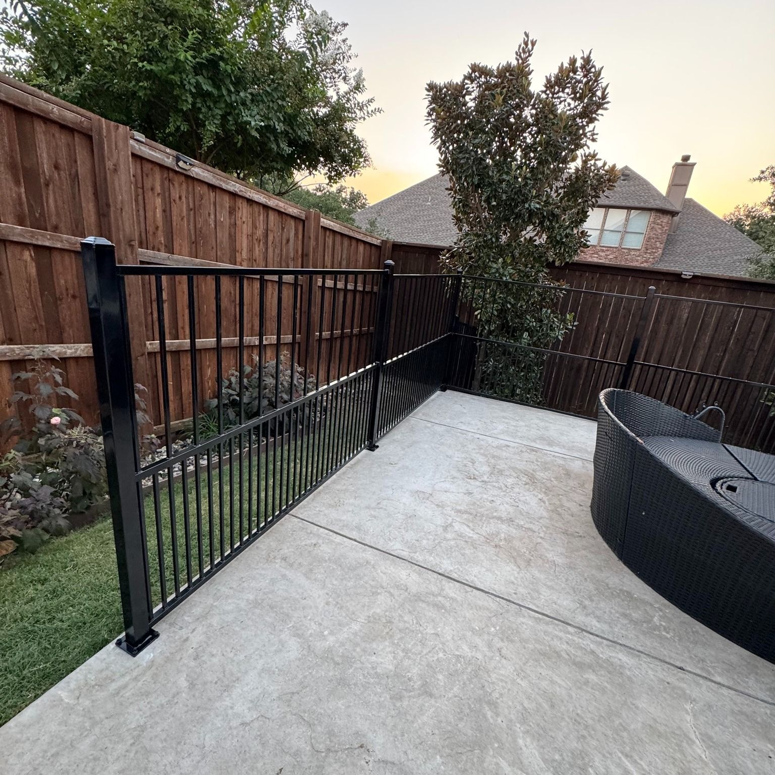 Black metal railing on a concrete patio next to a wooden fence, with a wicker sofa and greenery.