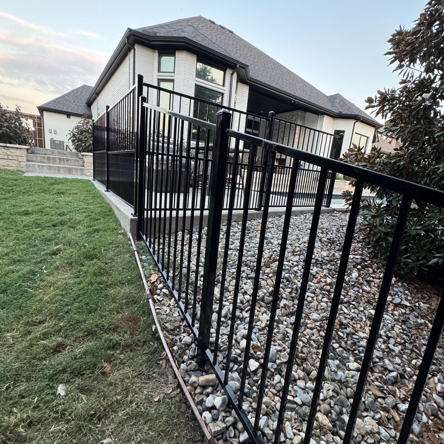 Black metal fence around a backyard with a stone bed and a white house.