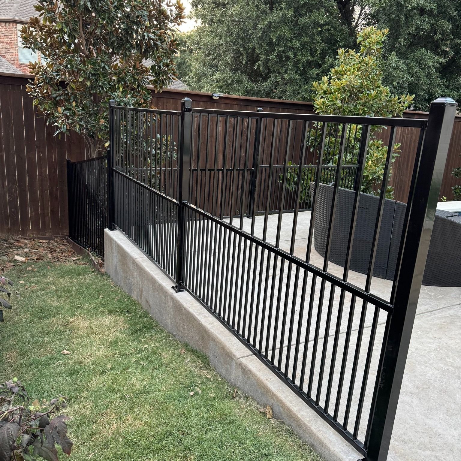 Black metal fence on a concrete wall separates a grassy area from a patio.