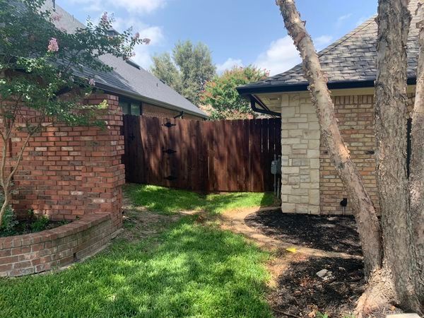 A narrow passageway between brick and stone buildings with a wooden fence, grass, and a tree.