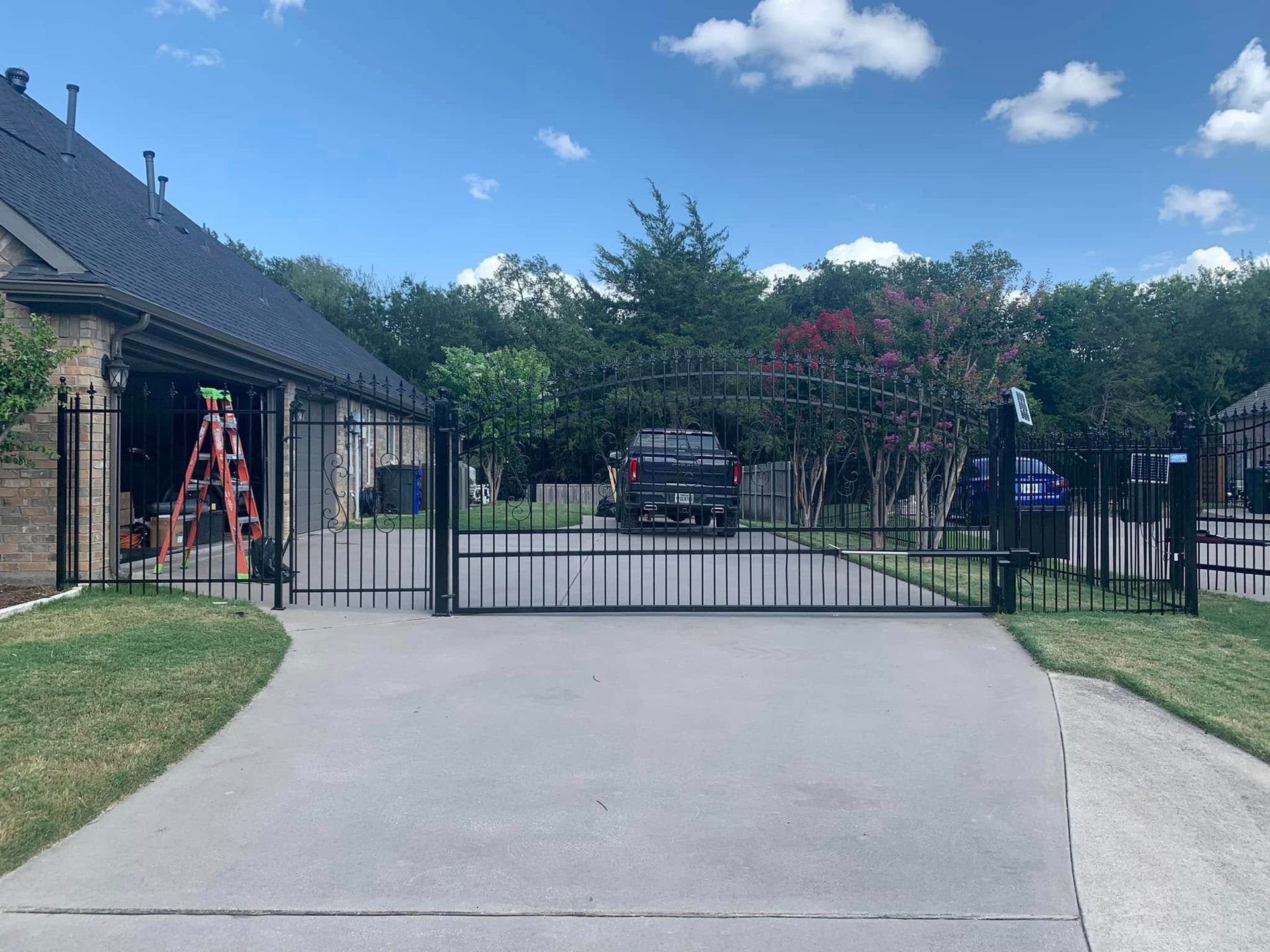 Black metal driveway gate with a concrete driveway and a house on a sunny day.