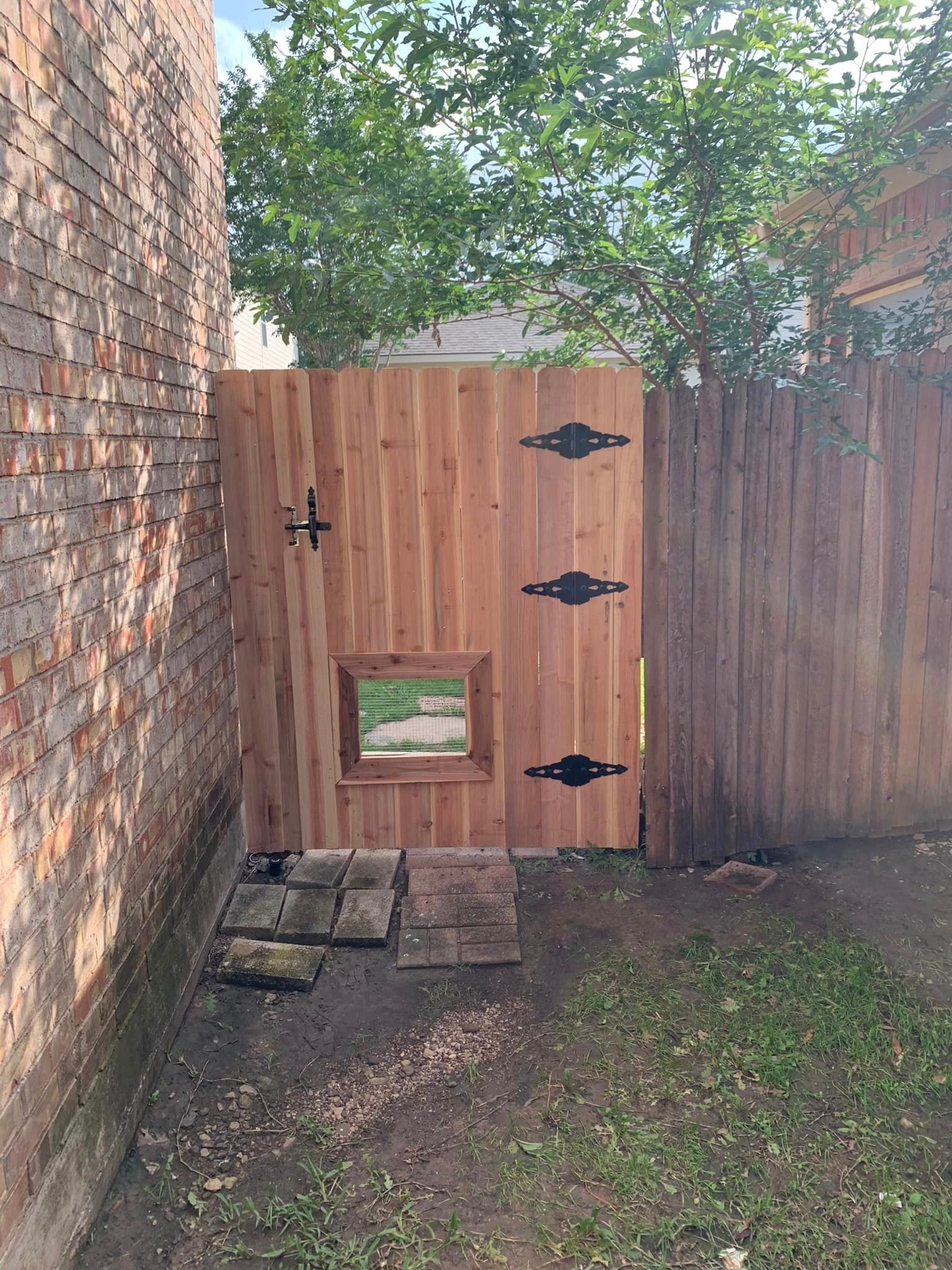 Wooden gate with window, black hinges, opening into grassy yard between brick walls.