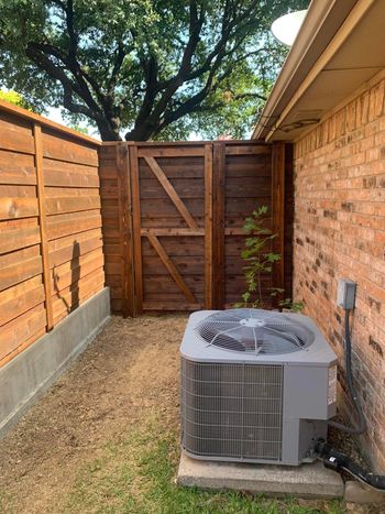 AC unit next to a brick wall. Wooden fence and gate in a gravel yard.