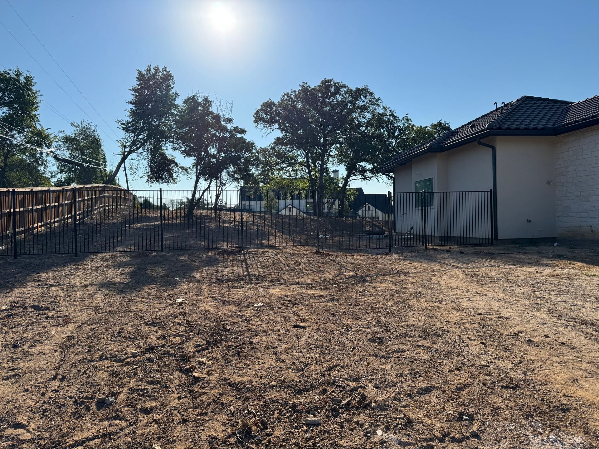 Barren lot with metal fencing, trees, and a building on a sunny day.