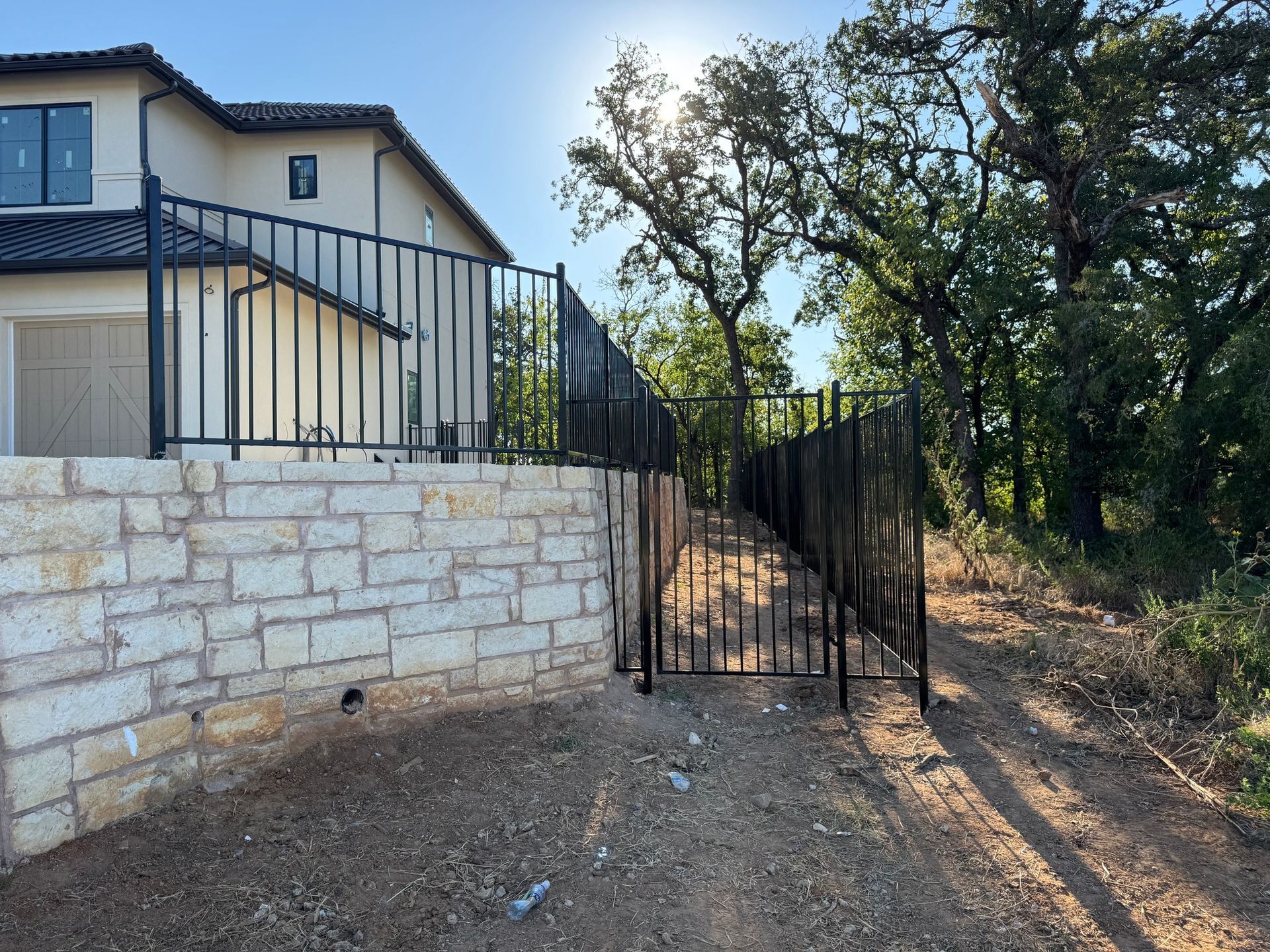 Black metal fence on a stone wall, leading to a path, next to a two-story beige house and trees.