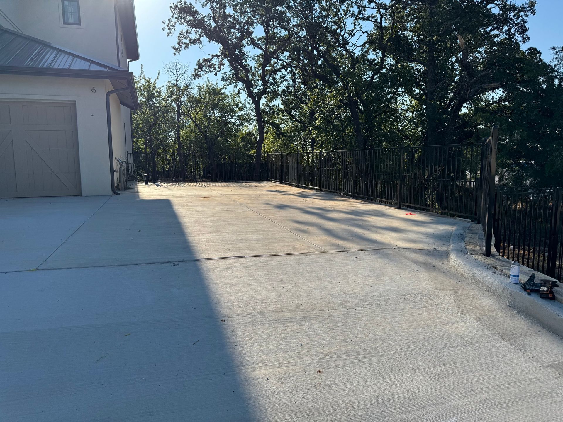 Concrete driveway next to a light-colored house with a garage. Trees and a black metal fence are in the background.