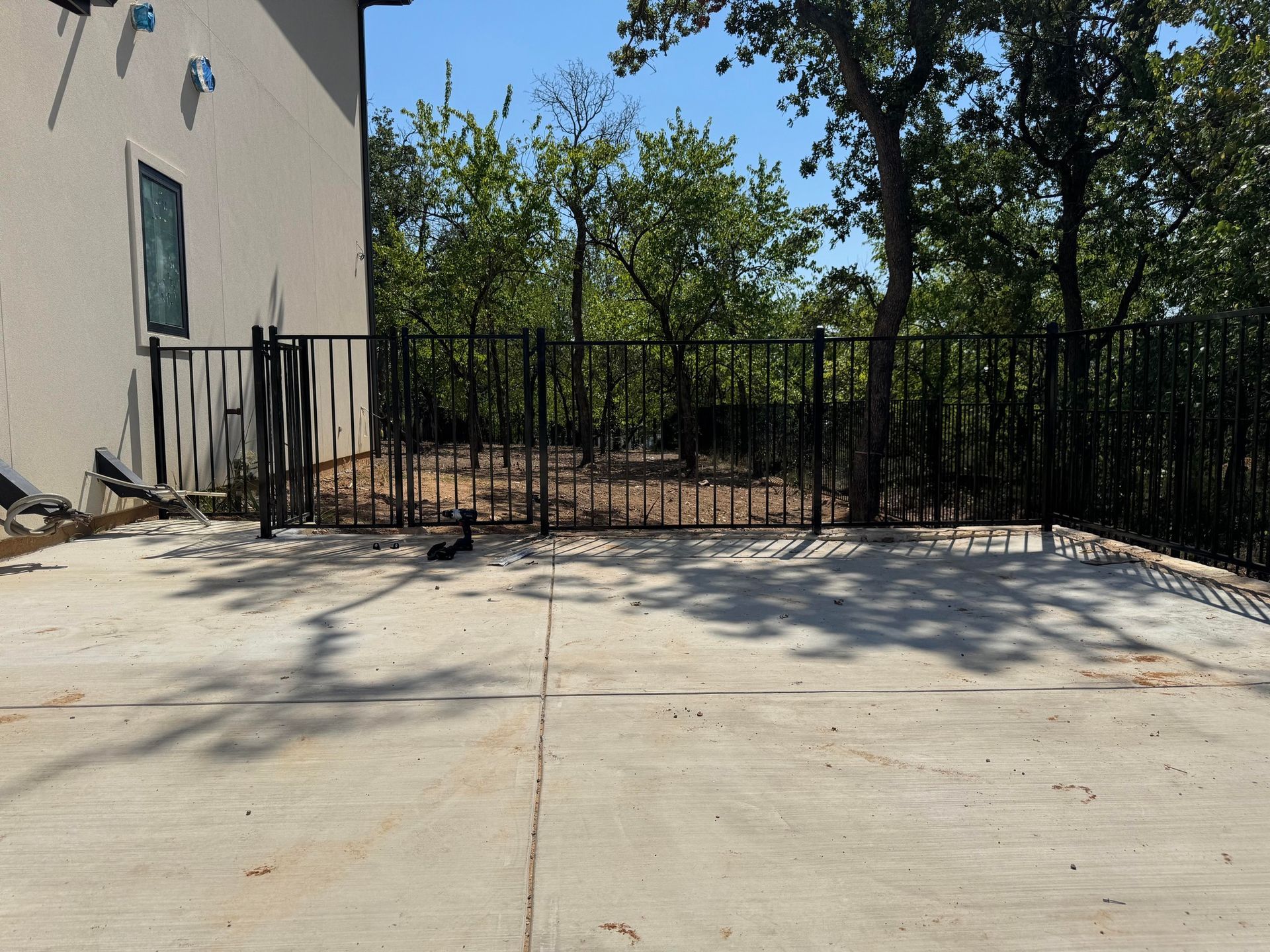 Concrete patio with a black metal fence and a building on the left; trees in the background.