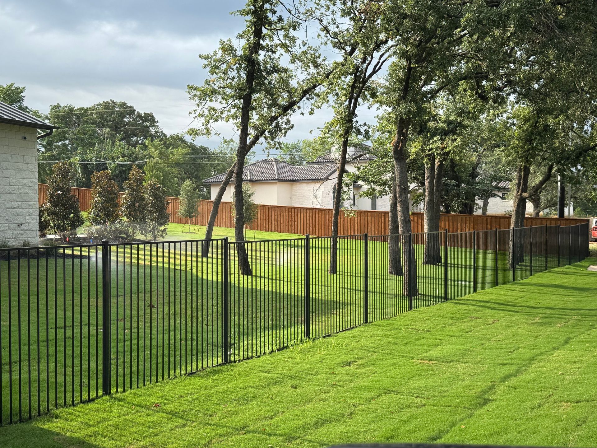 Black metal fence bordering a lush green lawn, trees in the background under a cloudy sky.