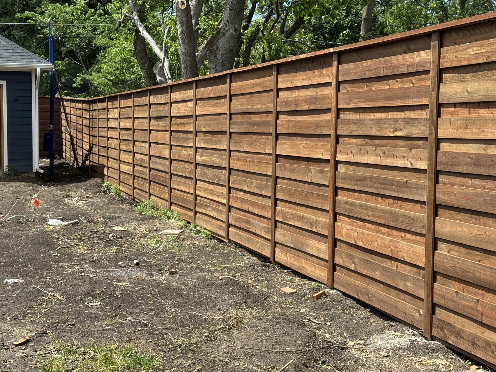 Brown horizontal slat fence in yard next to a blue building.