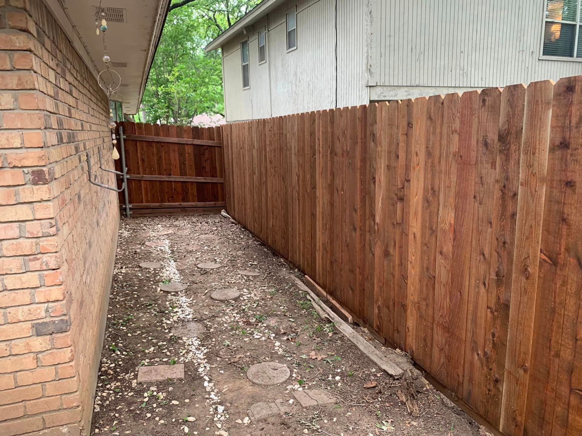 Narrow backyard with a wooden fence and brick wall. The ground has stepping stones and dirt.