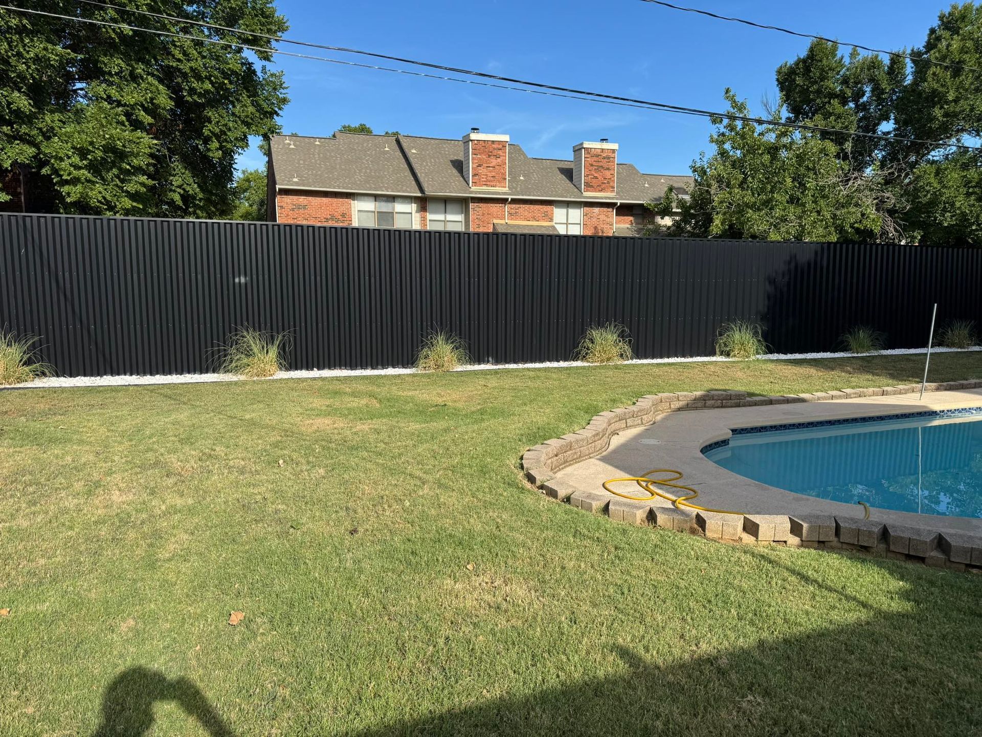 Black fence bordering a backyard with green grass, a pool, and a brick house in the background.