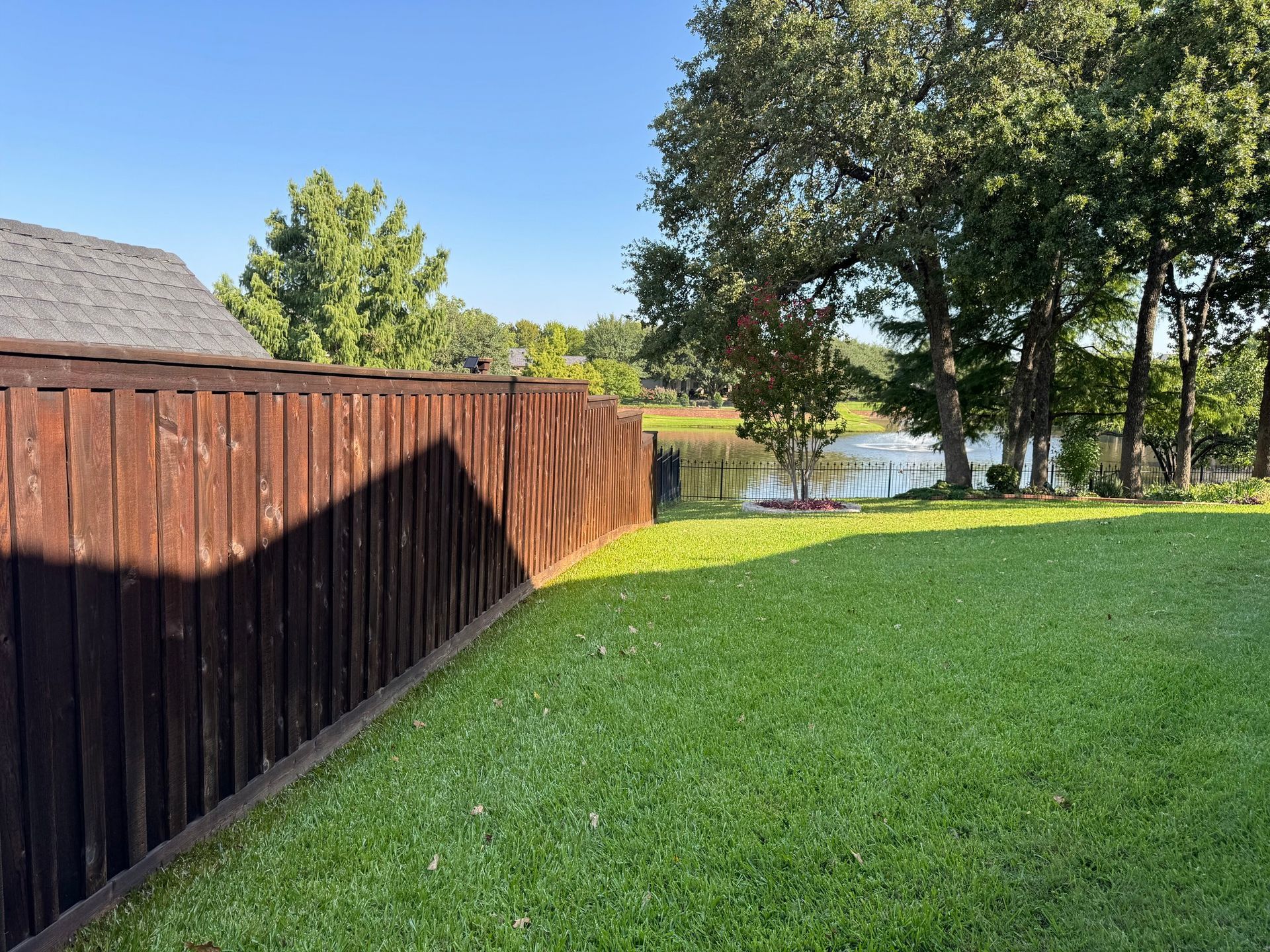 Wooden fence casting a shadow on green lawn, trees and lake in the background.