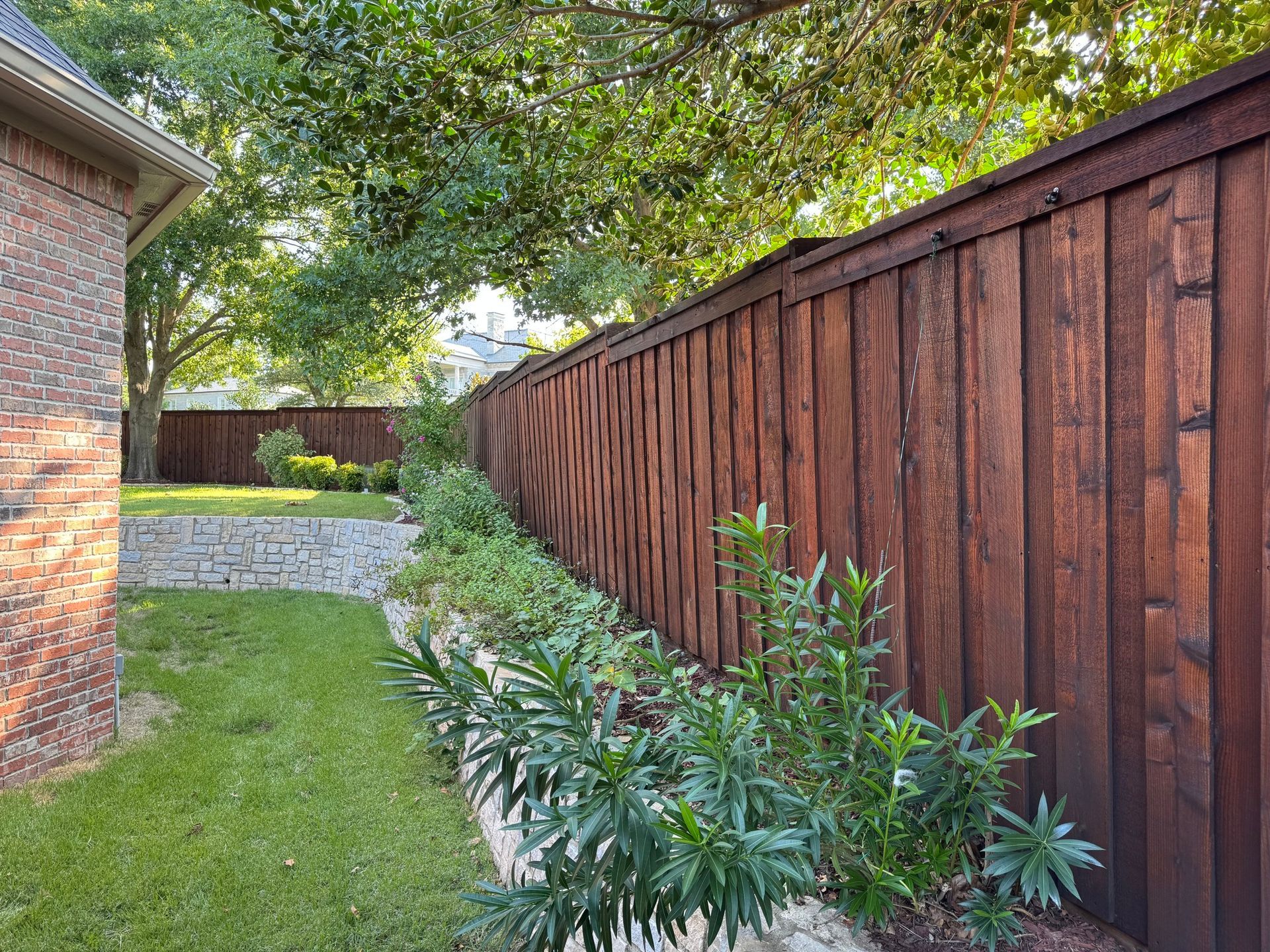Brown wooden fence borders a green lawn with a flower bed and a brick house in the background.
