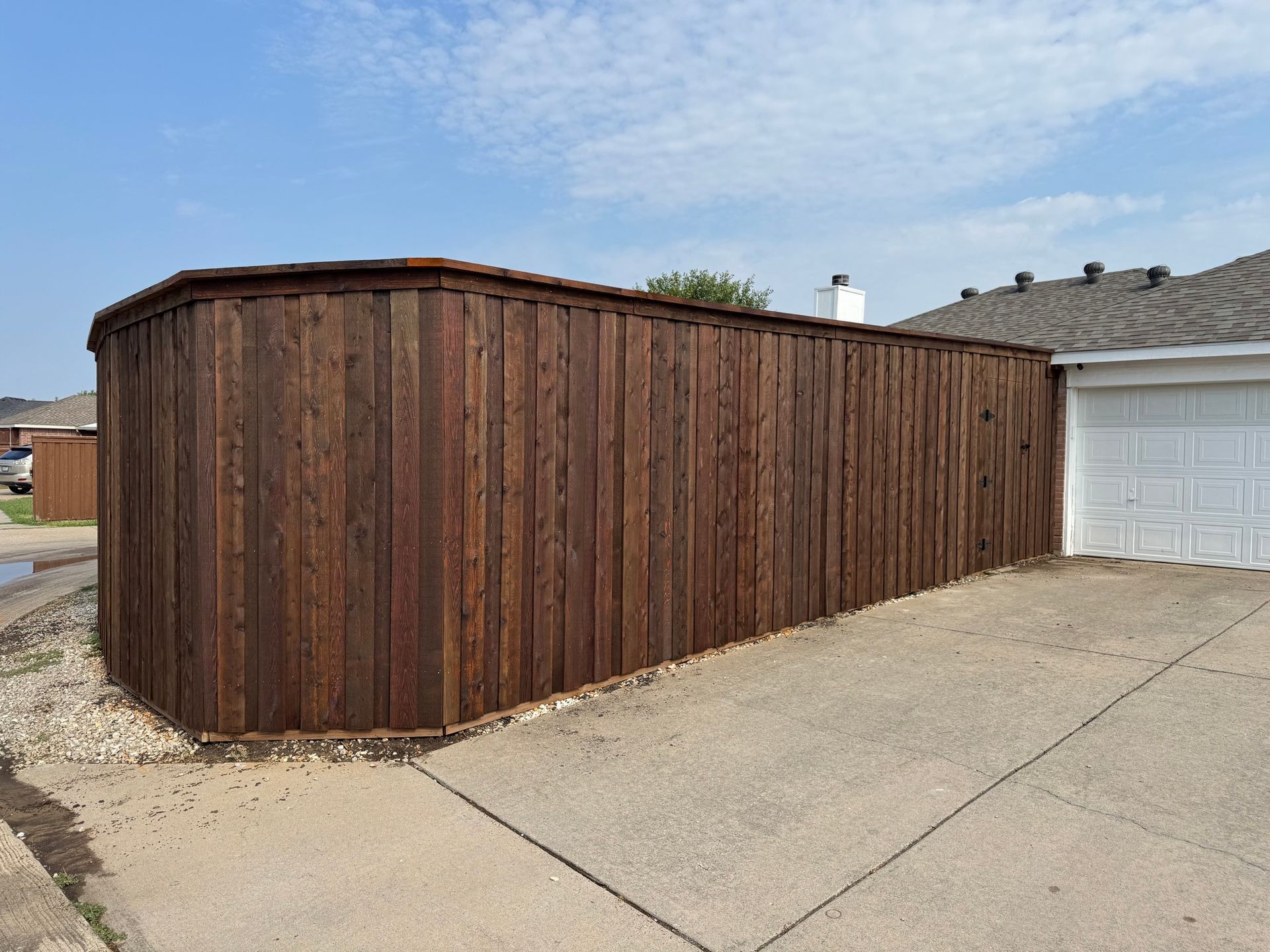 Dark brown wooden fence curving around a driveway next to a white garage on a sunny day.
