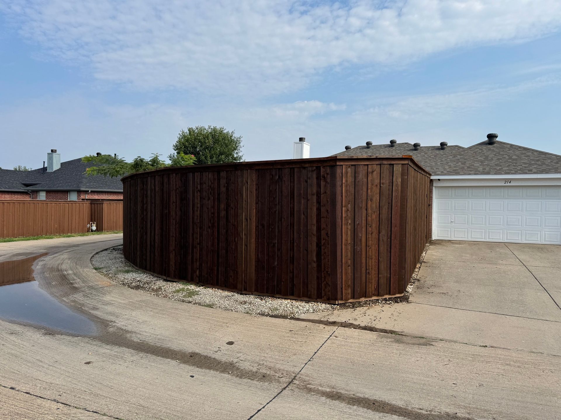 Wooden fence surrounding a gravel driveway, partially wet. Houses and a blue sky in the background.