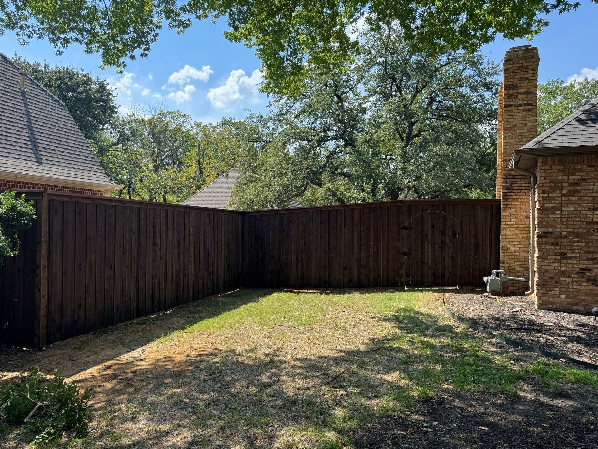 Brown wooden fence encloses a small backyard with patchy grass, trees, and brick house.