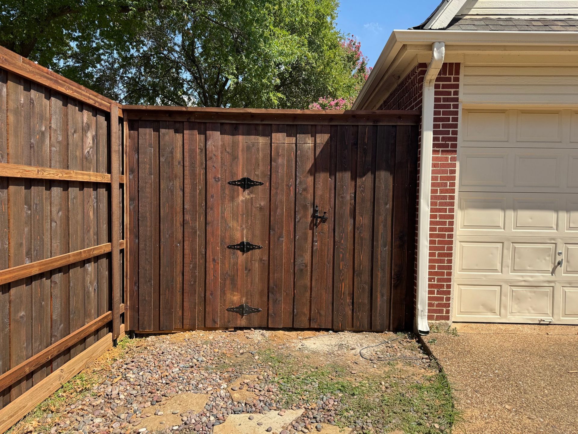 Brown wooden gate and fence beside a red brick garage and gravel driveway.