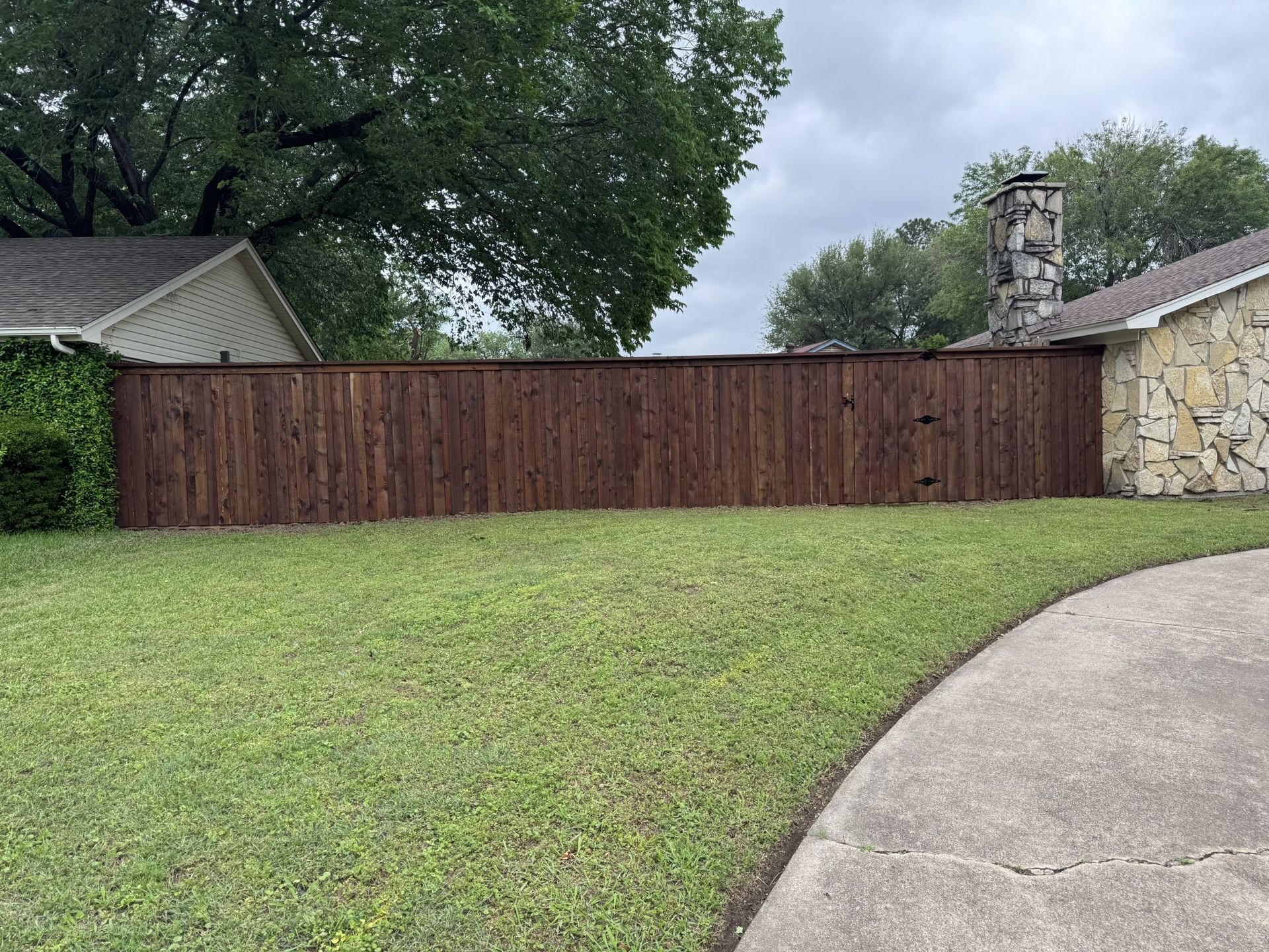 Brown wooden fence in front of a house with a green lawn and a cloudy sky.