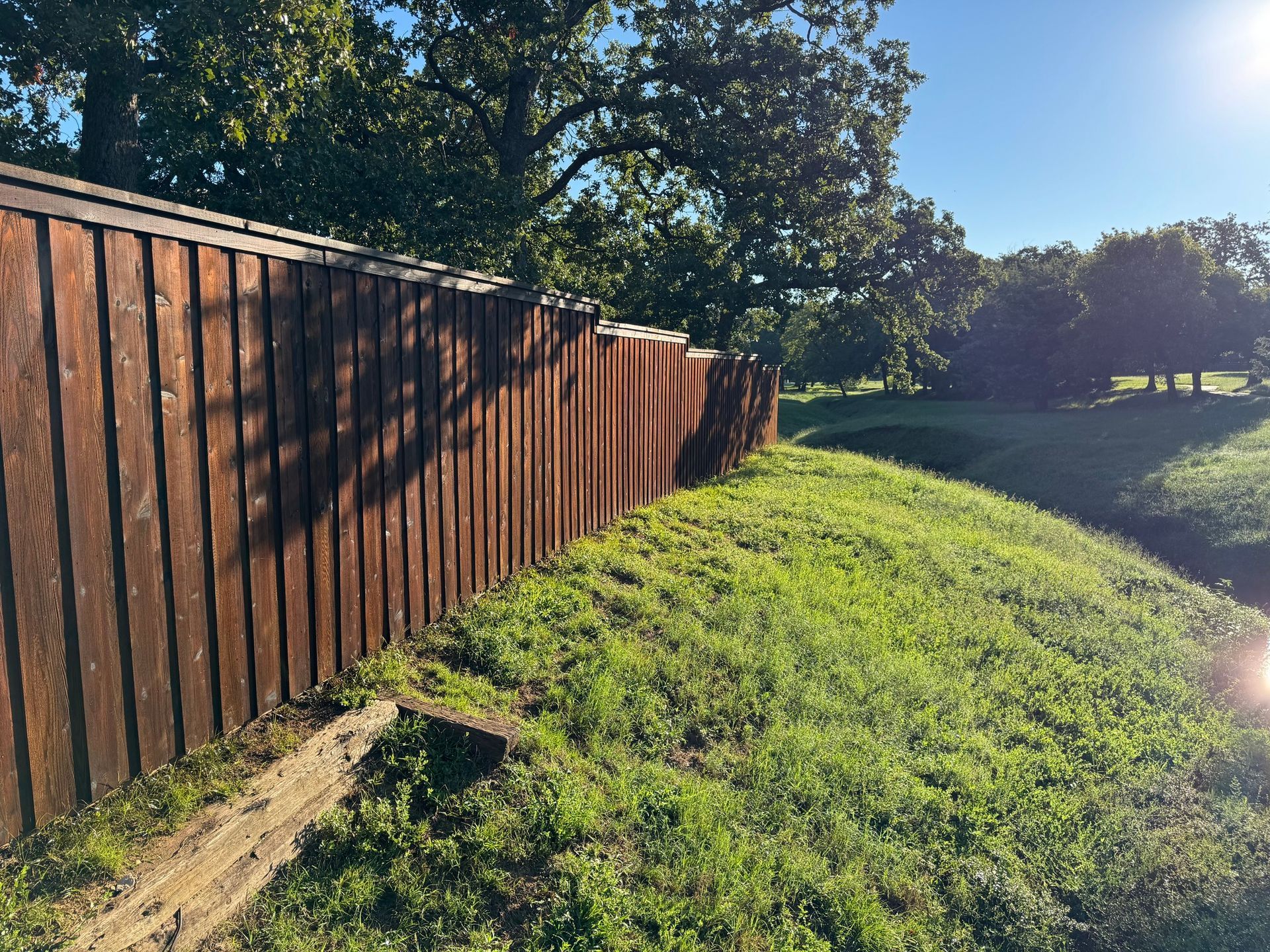 Brown wooden fence along a grassy hill in a park on a sunny day.