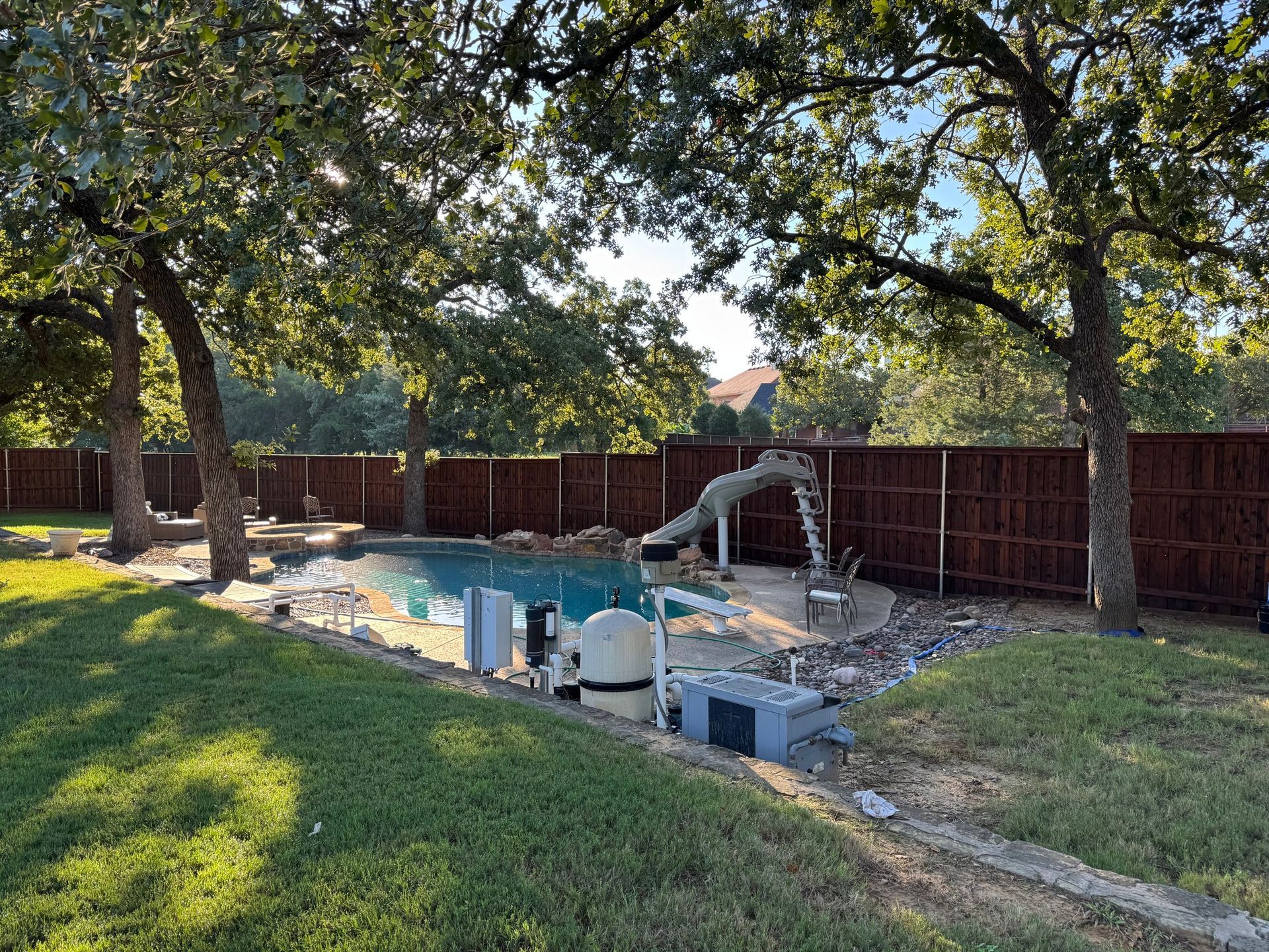 Backyard pool with slide, surrounded by grass and a brown wooden fence under trees on a sunny day.