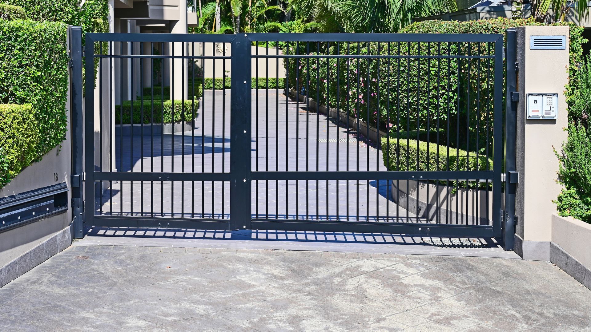 Black metal driveway gate; gray concrete; green hedges and lush foliage background.