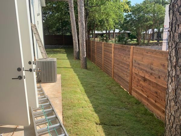 A newly installed wooden fence borders a grassy yard next to a house; a ladder leans against the wall.