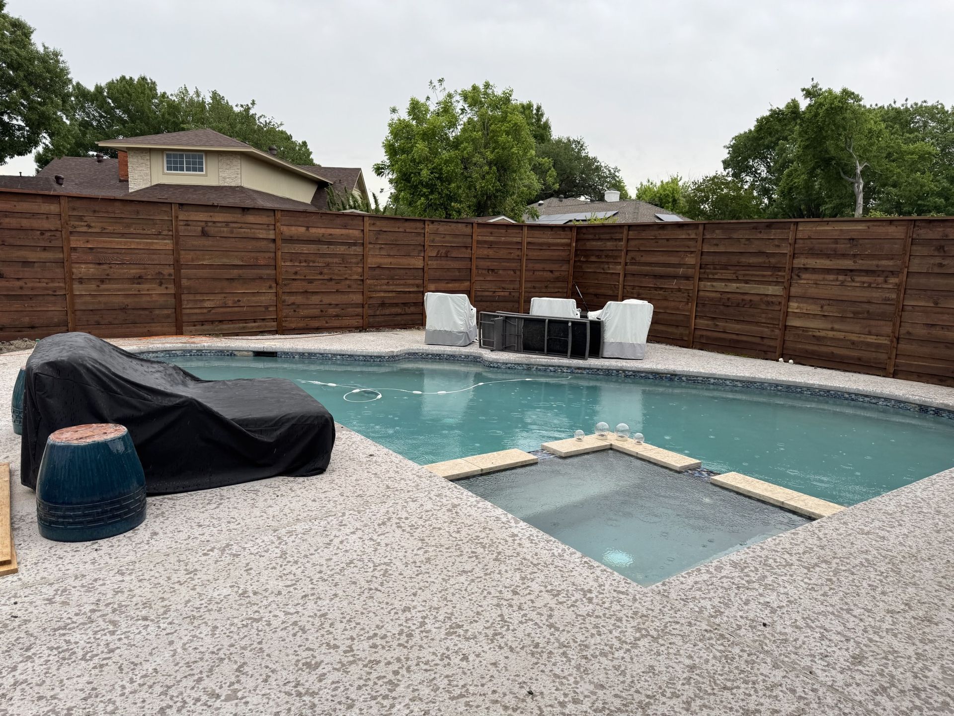 A backyard pool with a lounge chair, jacuzzi, and wood fence on a cloudy day.