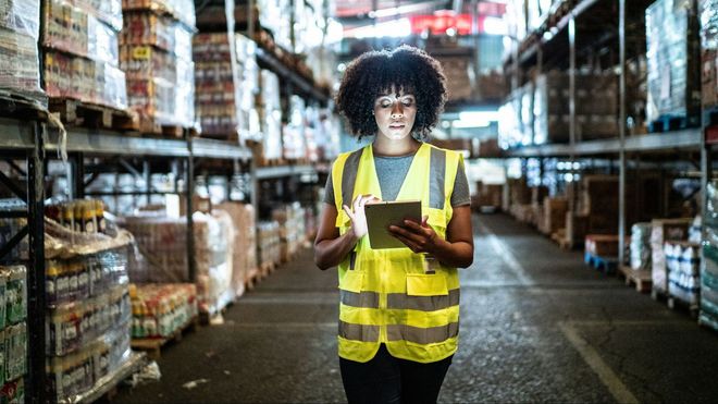 A woman in a yellow vest is using a tablet in a warehouse.