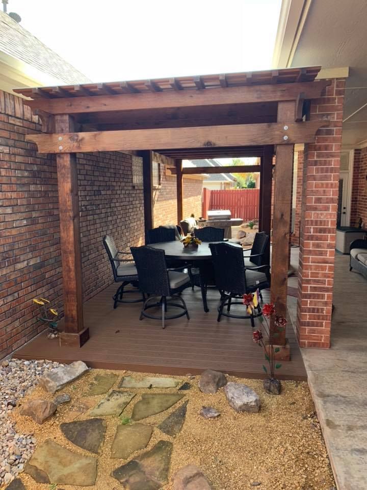 A wooden pergola over a deck with a round patio table and four chairs, situated between two brick walls.
