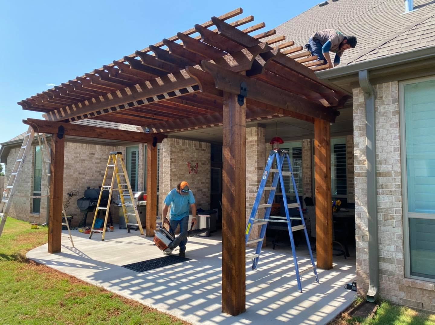 Workers construct a wooden pergola attached to a brick house, with ladders on a patio and one person on the roof.