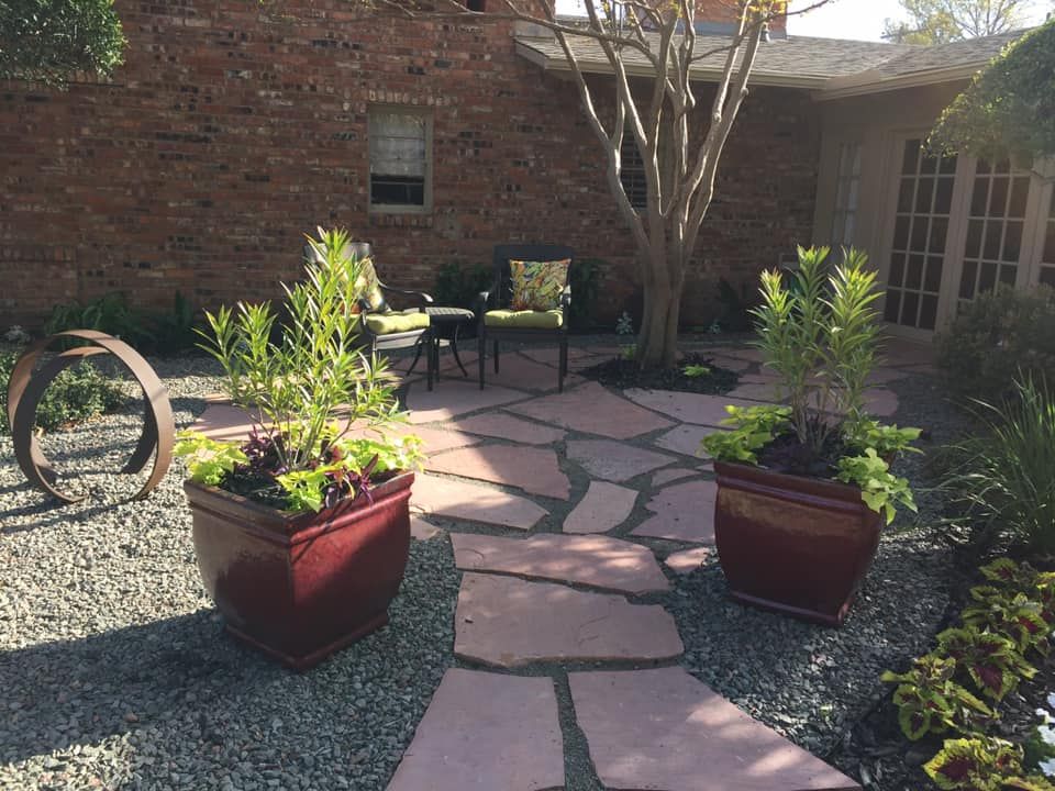 A stone patio with two dark red planters, a tree, and chairs in front of a brick building.