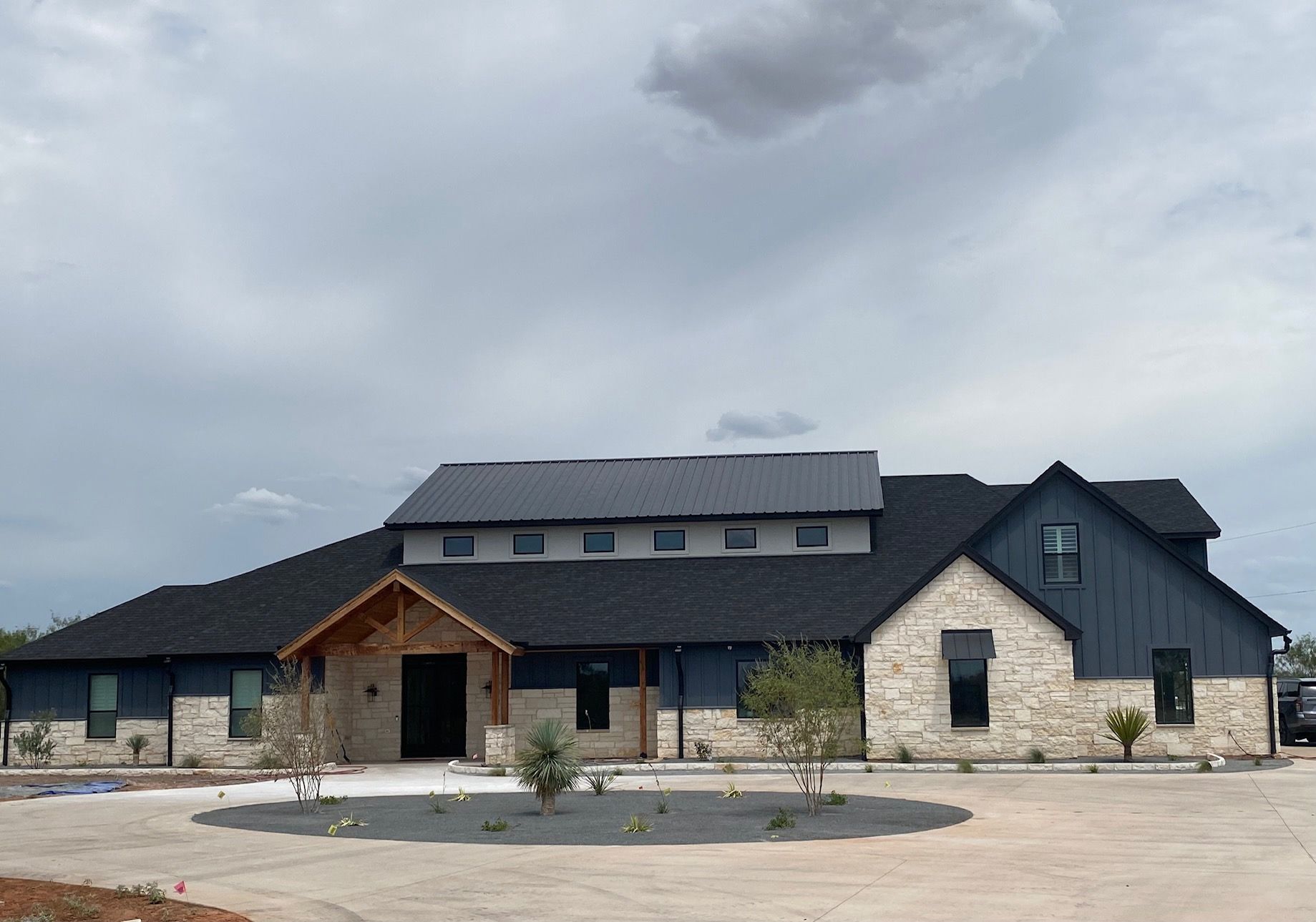 A large, modern house with dark roof shingles, dark blue vertical siding, and light stone exterior under a cloudy sky.
