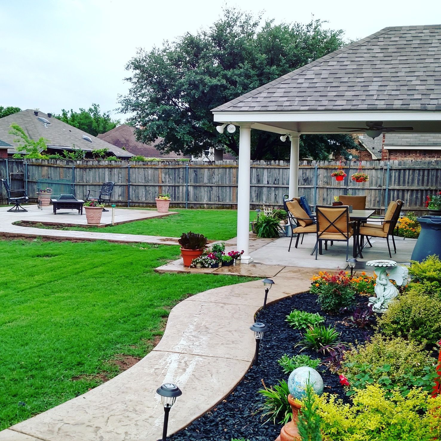 A landscaped backyard with a curved concrete path, a covered patio with outdoor furniture, and a grassy lawn.