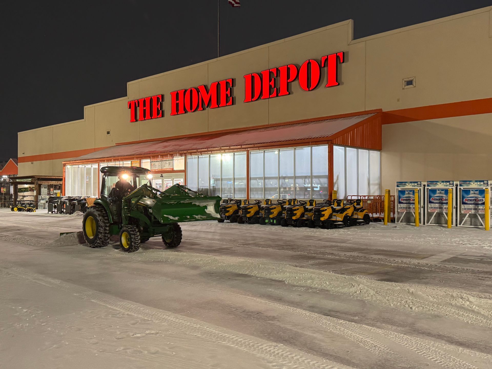 A green tractor with a front loader clears snow in front of a Home Depot store at night.