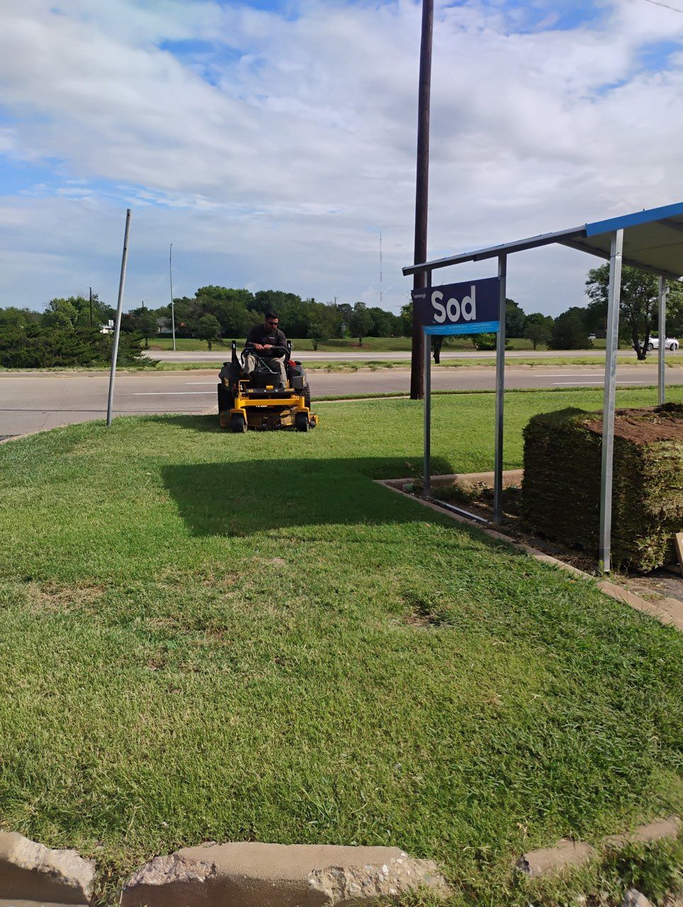 A person rides a yellow mower across a grassy lot next to a blue sign reading 