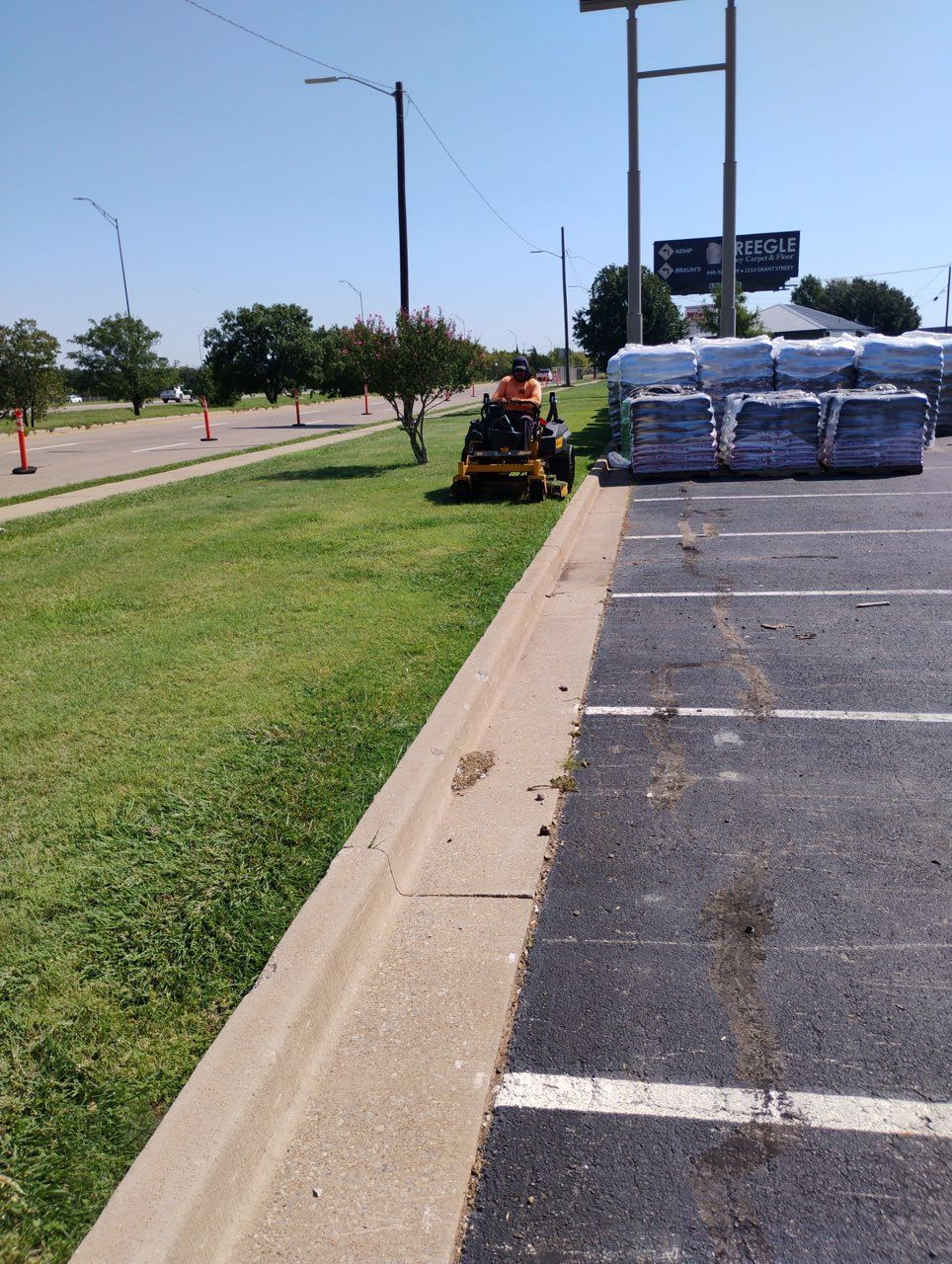 A worker mows the grass along a parking lot curb near a row of stacked, wrapped merchandise pallets under a clear sky.
